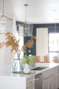 Loveliest Looks of Fall Home Tour Beautiful Fall Home Decor and Fall Fashion Ideas large white quartzite waterfall kitchen island with six gold barstools and a large green vase with yellow faux fall stems sitting on top of the countertop and gold kitchen lanterns hanging above the countertop then white kitchen cabinets with ann sacks patterned backsplash and gold hardware on the cabinets looking into a dark green dining room with a sputnik chandelier and burl wood dining room chairs