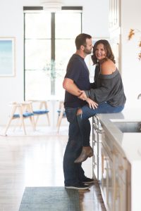 Loveliest Looks of Fall Home Tour Beautiful Fall Home Decor and Fall Fashion Ideas couple who design remodel and build homes together sitting in their kitchen hugging and laughing on a large quartzite waterfall countertop with white kitchen cabinets and gold lantern light fixtures