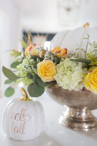 DIY Silver Urn Pumpkin Centerpiece for Fall silver urn with roses hydrangeas and eucalyptus with a ceramic pumpkin in the center sitting beside a hello fall pumpkin