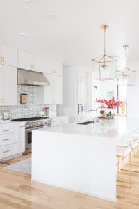 5 Tips to Decorate Your Home with Drew Barrymore Flower Home white quartz waterfall kitchen island with gold bar stools and ann sacks patterned honeycomb backsplash with gold kitchen lanterns and pink flowers styled in a kitchen