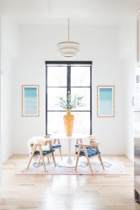5 Tips to Decorate Your Home with Drew Barrymore Flower Home yellow floor and table vase with faux olive branches sitting on a white tulip table with vintage wishbone chairs in a room with white walls and black windows