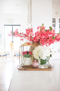 5 Tips to Decorate Your Home with Drew Barrymore Flower Home white quartz waterfall kitchen island with gold bar stools and ann sacks patterned honeycomb backsplash with gold kitchen lanterns and pink flowers styled in a kitchen